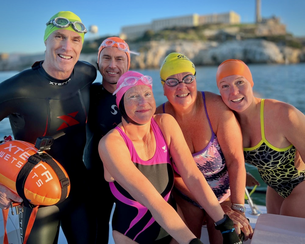 A group of smiling swimmers preparing for an Alcatraz swim in San Francisco Bay, with wetsuit and skin swimmers ready for the challenge.