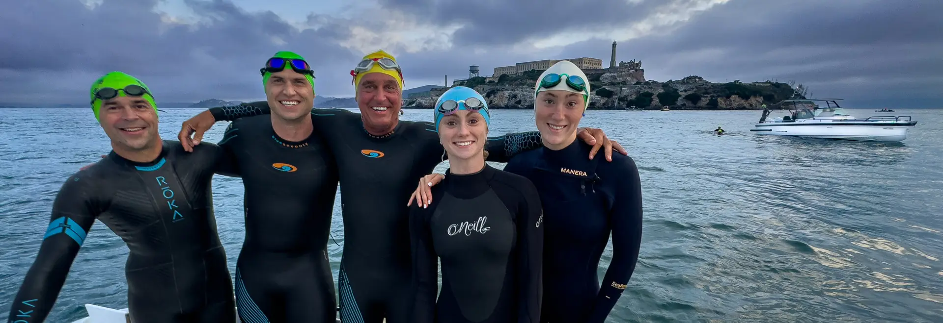 Five smiling swimmers, some in wetsuits and some skin, preparing to jump into the water at Alcatraz for a swim to San Francisco.